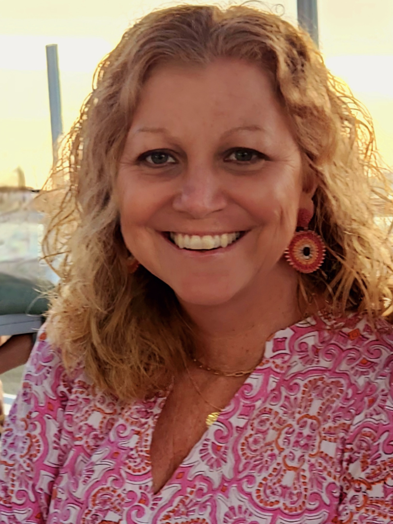 A smiling woman with curly blonde hair wearing a pink and white patterned blouse and colorful earrings.