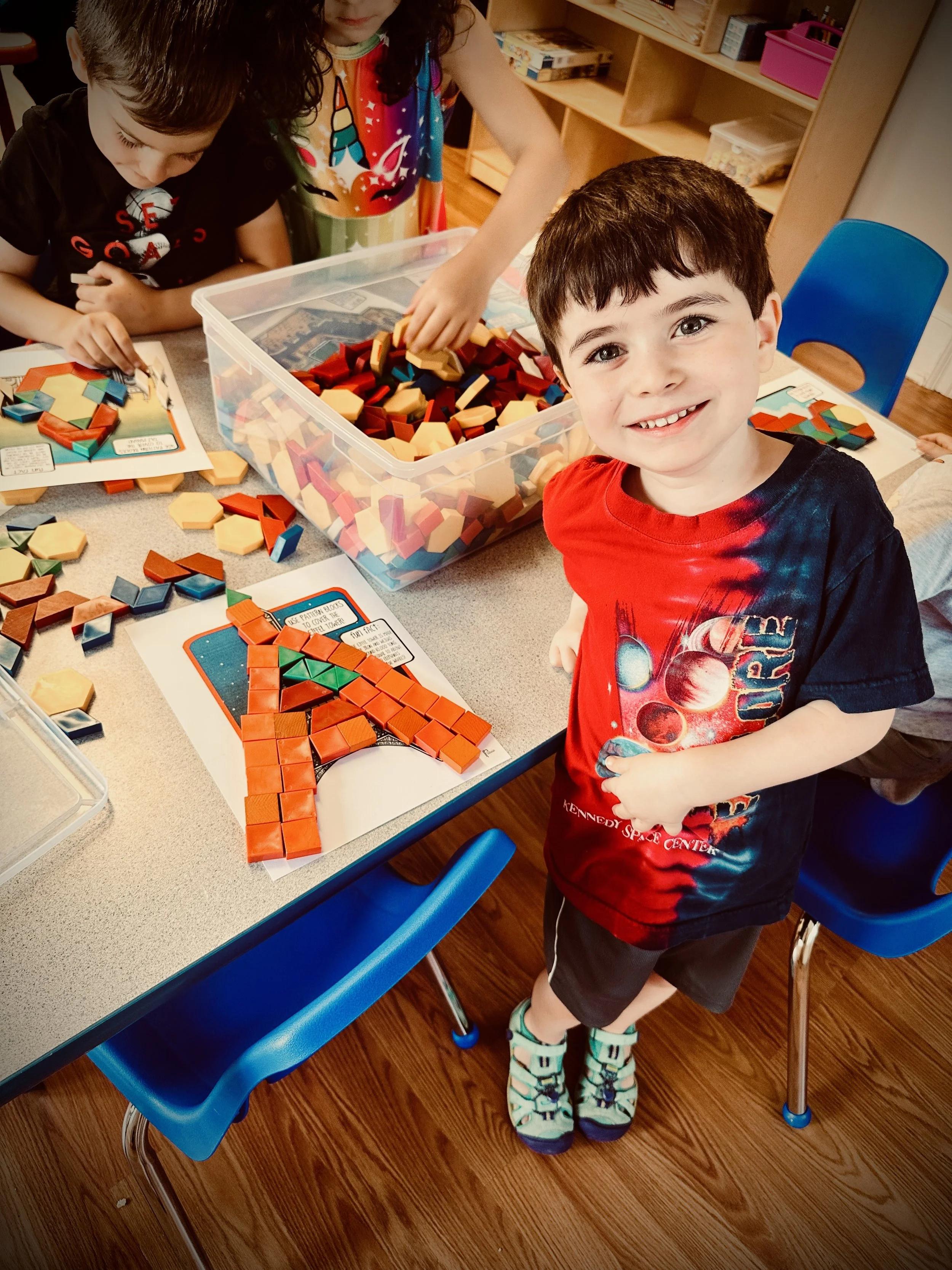A young boy with dark hair and a red and black tie-dye shirt stands at a table playing with colorful geometric toys. Two girls are visible in the background also engaged with toys. The table has a plastic container filled with red, yellow, and blue foam blocks, and there are templates or worksheets on the table. The setting appears to be a classroom or a playroom with wooden floors and shelves in the background.