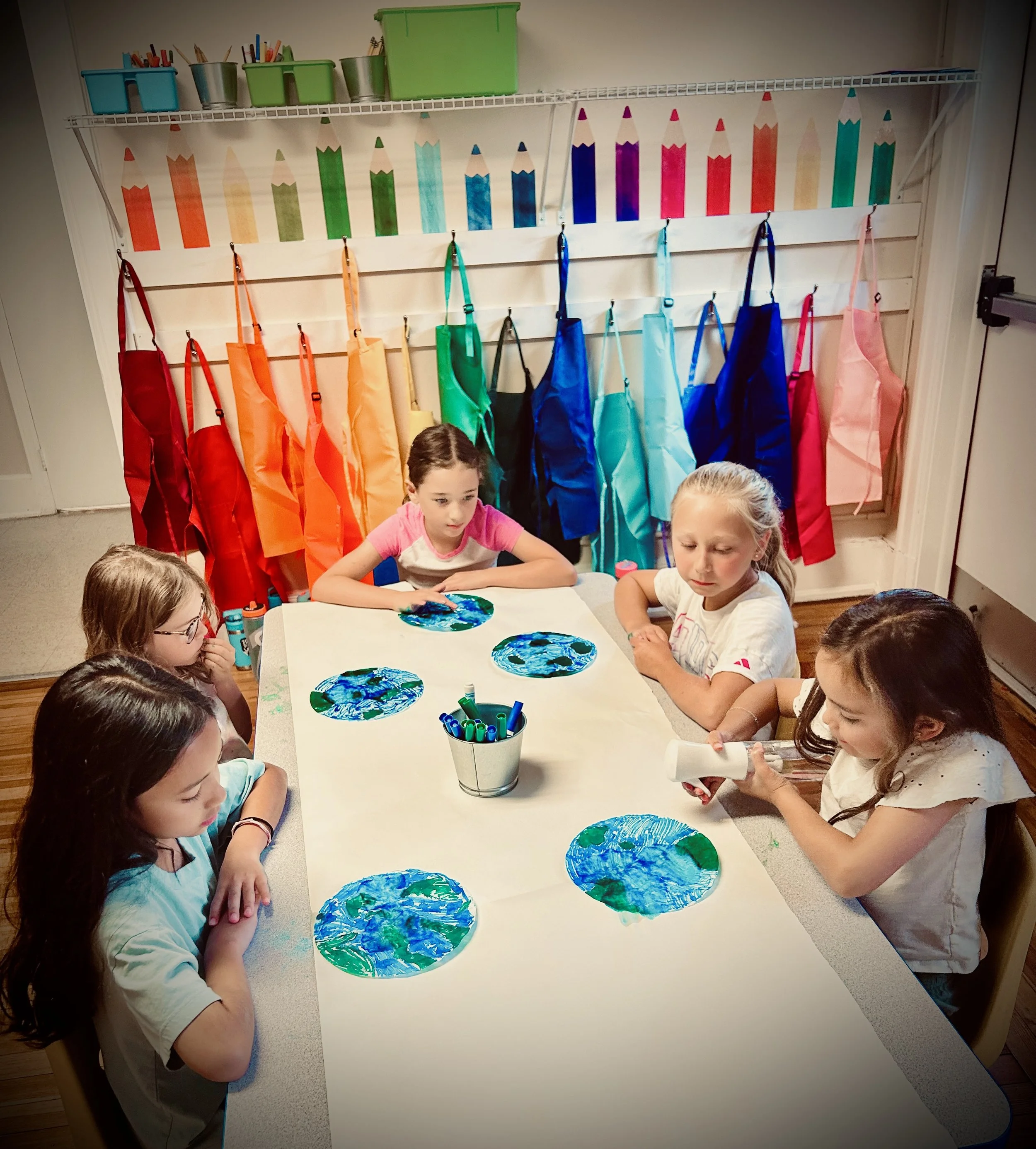 Children engaged in a painting activity at a table, with colorful painted paper plates in front of them. There are hanging aprons in various bright colors and a wall decorated with large, colorful pencil illustrations.