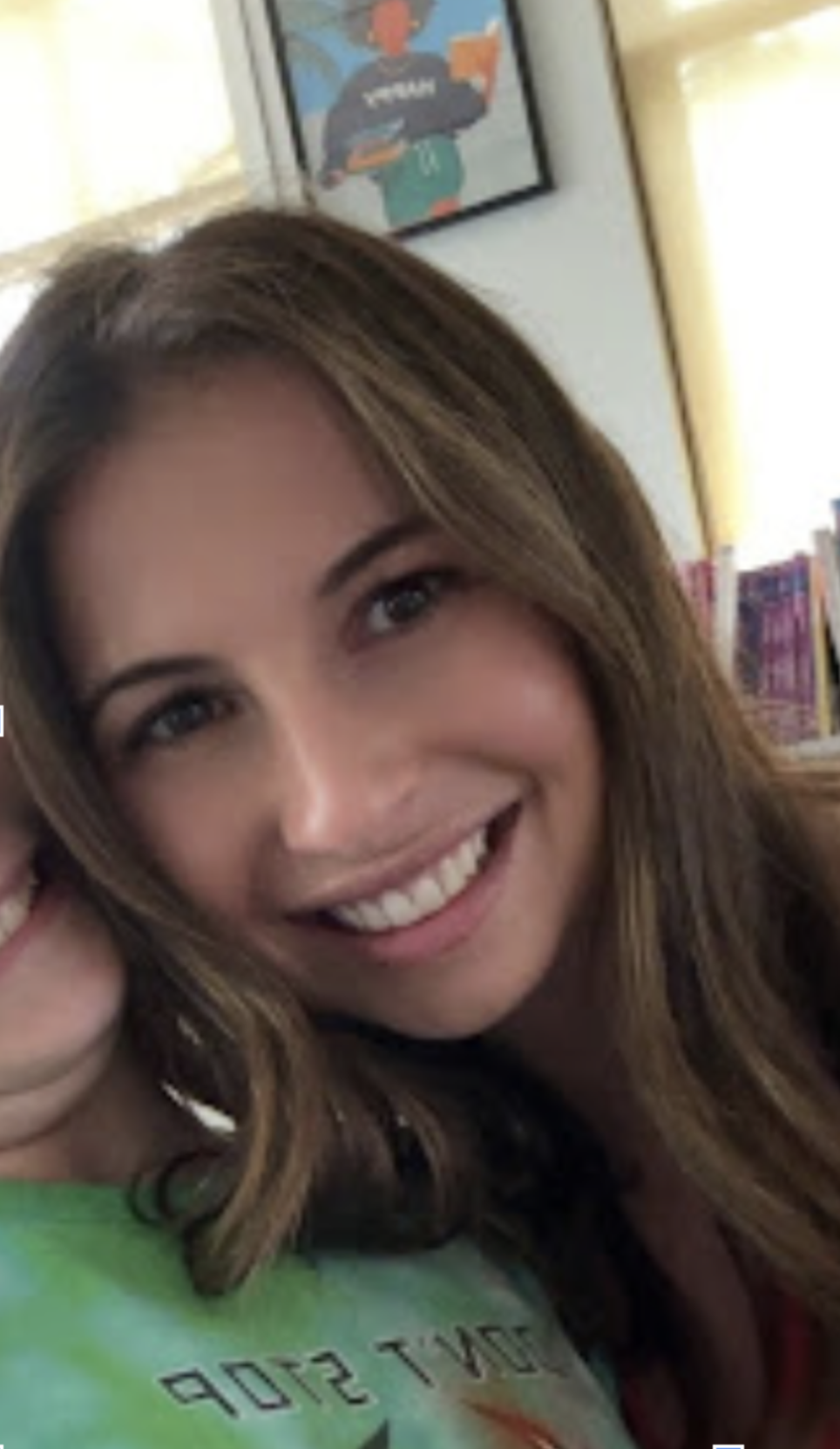 A smiling woman with long, wavy brown hair, posing indoors in front of bookshelves and a framed illustration of a person holding a book.