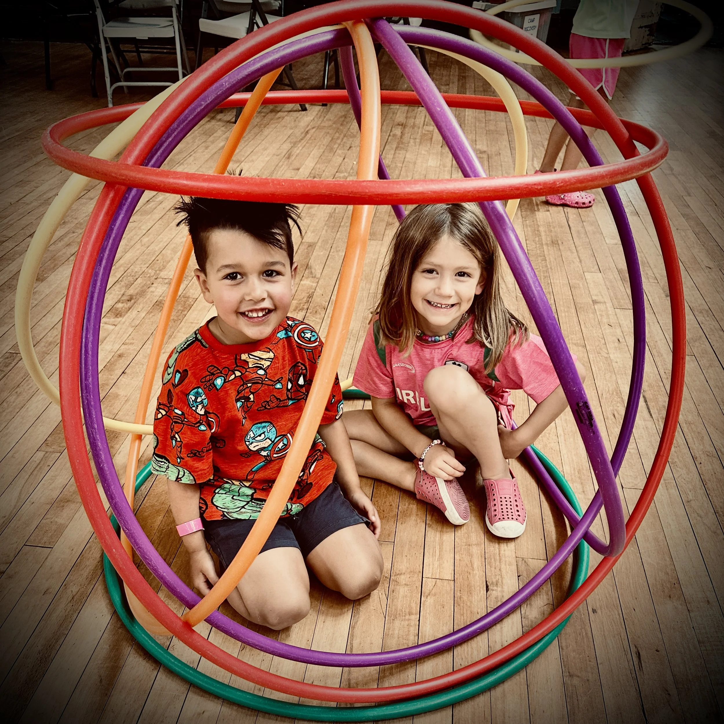 Two children sitting inside a colorful, spherical climbing structure made of interconnected hoops in purple, red, orange, yellow, and green, on a wooden floor.