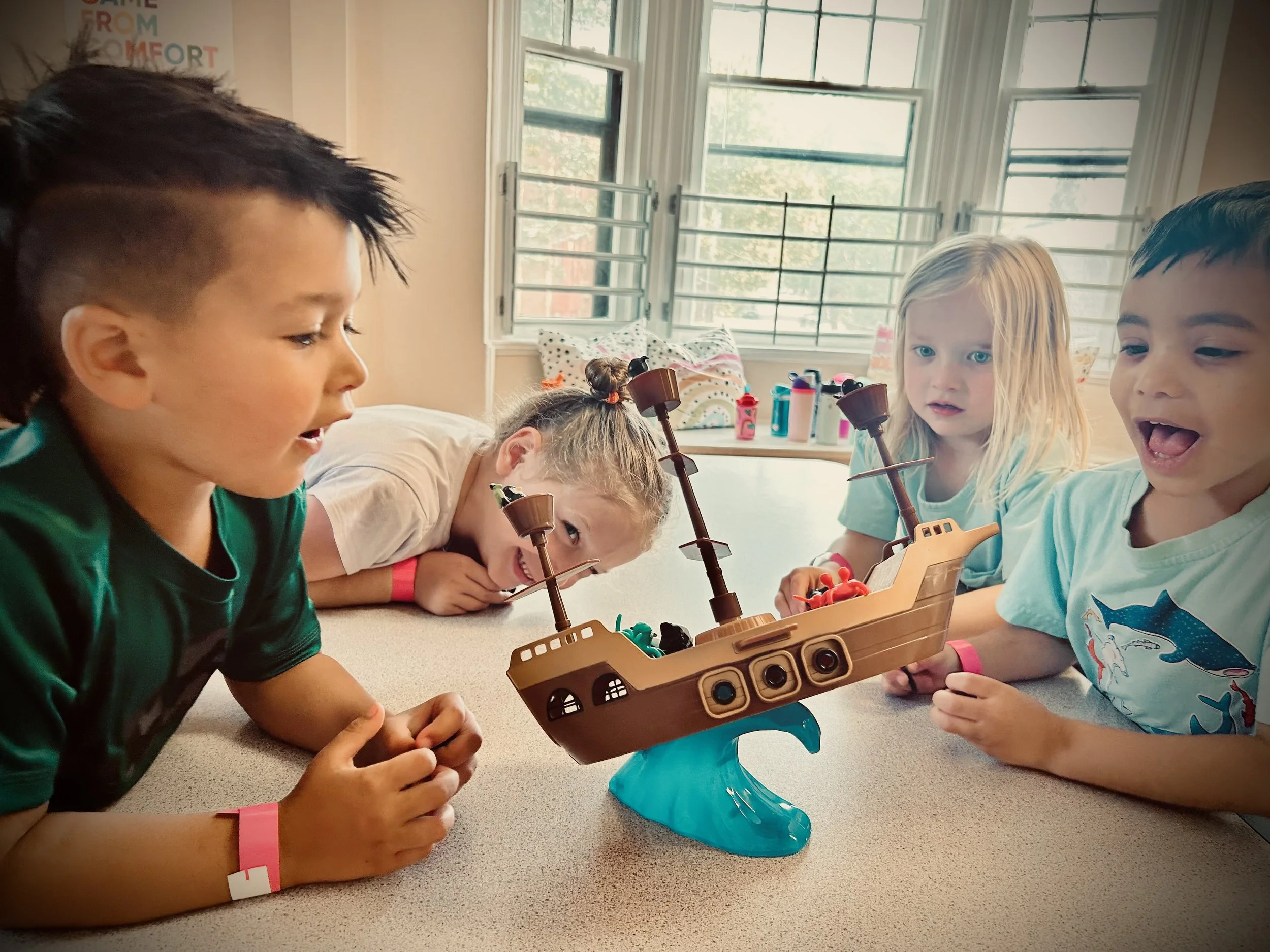 Group of children playing a board game together in a brightly lit room with large windows.