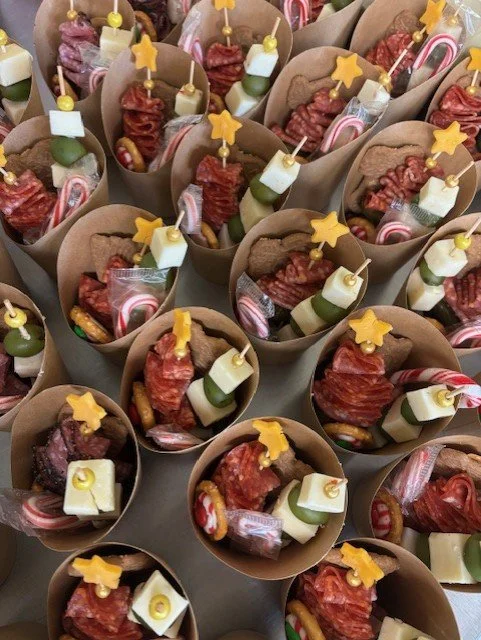 Individual cups filled with assorted snacks including bacon strips, cheese cubes, pickles, and candy canes, topped with yellow star-shaped decorations.