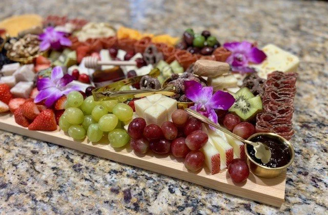 Charcuterie board with cheese, grapes, fruit, crackers, and flowers on a granite countertop.