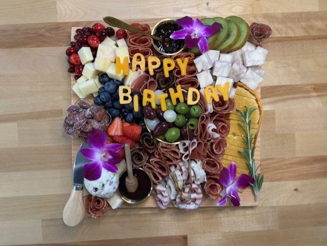 Charcuterie board with assorted fruits, cheeses, meats, and crackers, decorated with purple orchids spelling out "Happy Birthday" on a wooden table.