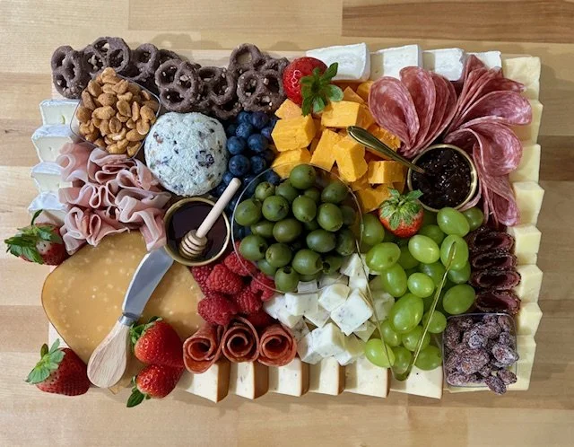 A cheese and charcuterie board with grapes, strawberries, blueberries, chocolate-covered pretzels, cheese cubes, slices of deli meats, honey, jam, and crackers.
