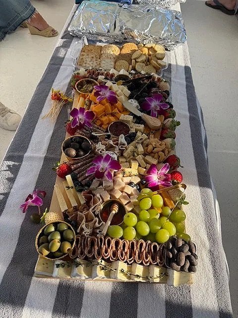 A cheese platter with green grapes, black olives, and purple orchids, set on a table with foil-wrapped items at the top.