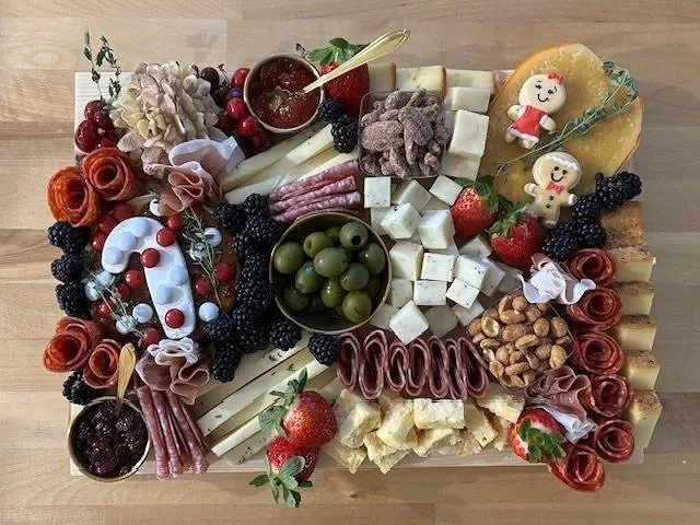 Charcuterie cheese board with strawberries, blackberries, green olives, strawberries, prosciutto, cheese cubes, nuts, fruit slices, and decorative baby figurines on a wooden surface.