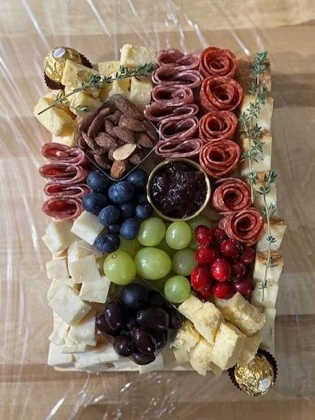Assorted cheese platter with grapes, blueberries, red and green currants, sliced cured meats, and a small jar of jam.