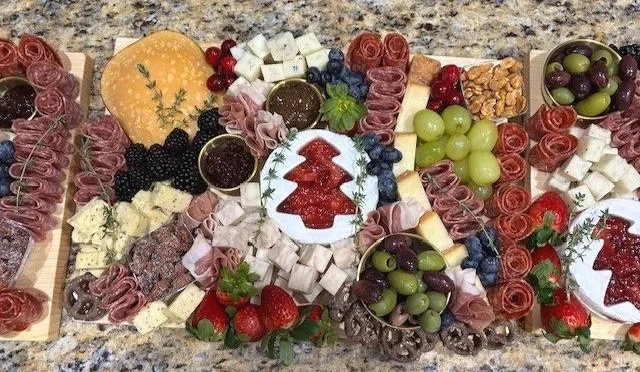 Assorted cheeses, grapes, strawberries, blueberries, blackberries, sliced meats, honey, and holiday-themed snacks on a granite countertop.
