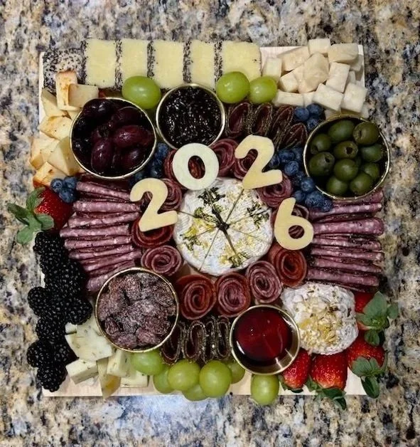 Chocolate cake with white frosting and gold accents, surrounded by grapes, strawberries, blueberries, blackberries, cheese, cured meats, jelly, and cookies arranged in the shape of the year 2026.