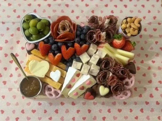 A cheese and fruit platter with strawberries, blueberries, grapes, heart-shaped cheese, sliced melon, and chocolate roses, arranged on a pink tablecloth with small hearts.