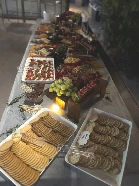 A long table with a variety of snacks, including crackers, grapes, cheese, fruit, and other finger foods, set up for a gathering or party at night.
