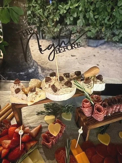 A dessert display with various treats, including cheesecake with chocolate chunks, strawberries, and cheese cubes, decorated with greenery and a 'light before' cake topper.