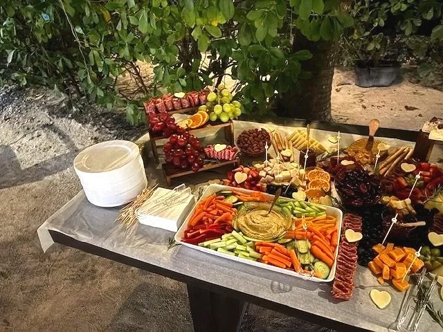 Charcuterie and vegetable platter with grapes, cheese, crackers, and dips on a black table outdoors, surrounded by bushes.