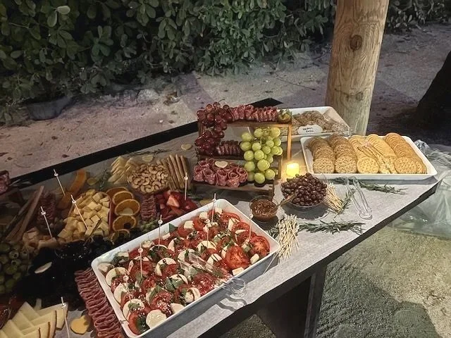 Outdoor buffet table with various cheeses, grapes, sliced meats, crackers, and a tray of caprese salad skewers, set against a background of greenery and a wooden post