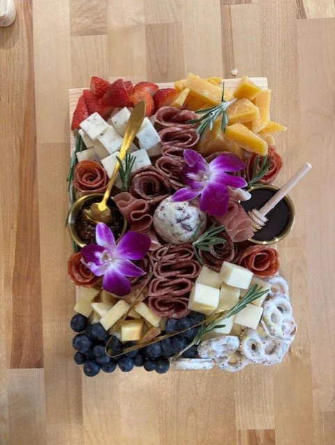 Charcuterie board featuring sliced meats, cheeses, strawberries, blueberries, crackers, olives, honey, and purple flowers on a wooden surface.