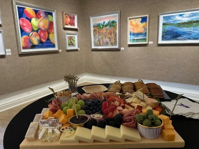 A cheese and fruit platter with grapes, berries, slices of cheese, crackers, and assorted meats on a black table in an art gallery with colorful paintings on the wall.
