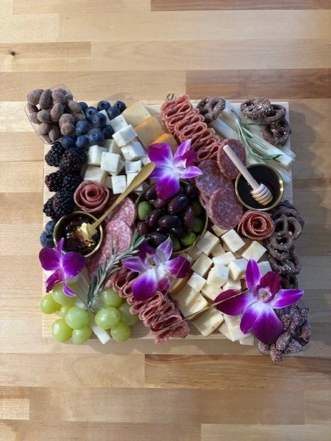 Cheese and charcuterie platter with grapes, blueberries, blackberries, salami, cheese cubes, cheese slices, purple and green grapes, edible flowers, honey jars with honey dippers, and rosemary sprigs on a wooden surface.
