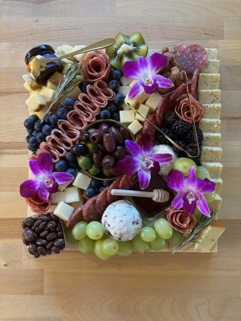 A cheese and charcuterie board with grapes, blueberries, sliced meats, cheese cubes, decorative flowers, olives, kiwi, and crackers.