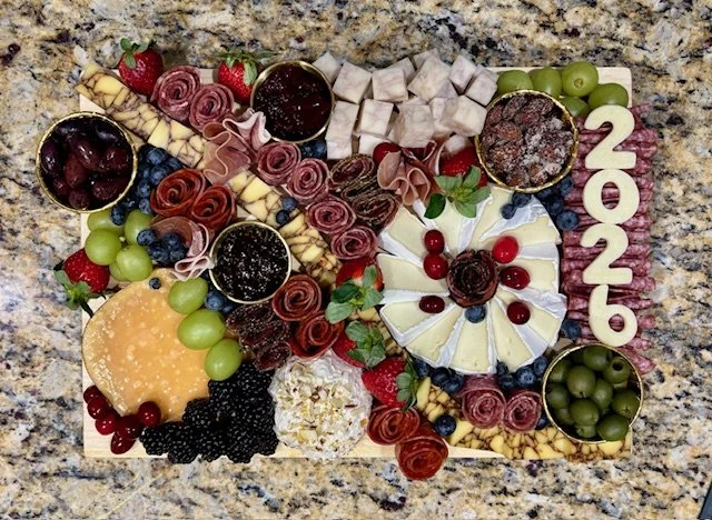 A charcuterie board with cheeses, grapes, berries, crackers, and jams, decorated with the year 2026 made of pink and white letters.