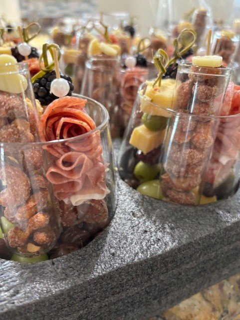 Multiple glasses filled with assorted charcuterie and fruit skewers, including slices of meats, cheeses, grapes, and other snacks, arranged on a gray stone surface.