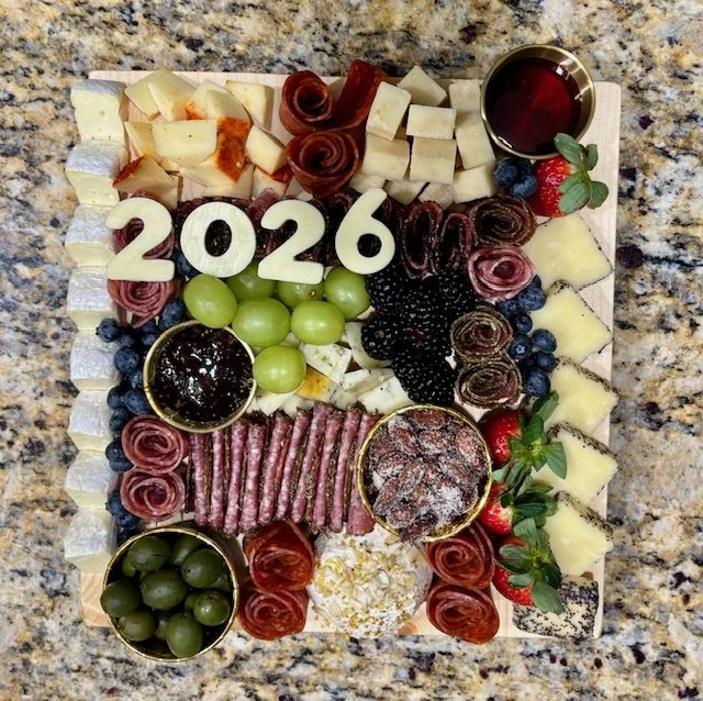 Charcuterie board with cheese, grapes, berries, cured meats, honey, and the numbers '2026' in the center, on a granite countertop.