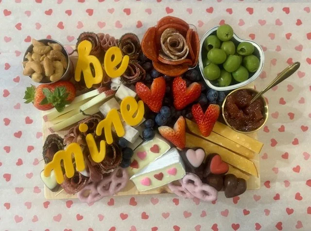  assortment of sweets, strawberries, green grapes, and cheese on a heart-patterned tablecloth, spelling out 'be mine' in yellow decoration.