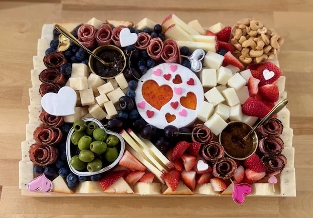 A cheese and fruit platter decorated with strawberries, blueberries, green grapes, nuts, and heart-shaped chocolates. There are two small bowls with jam, a round cheese with heart-shaped cutouts, and decorative heart and flower-shaped paper accents.