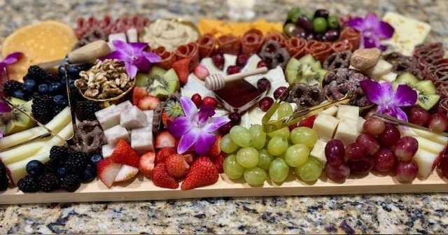 Charcuterie board with grapes, strawberries, blueberries, cheese, chocolate, nuts, and flowers.
