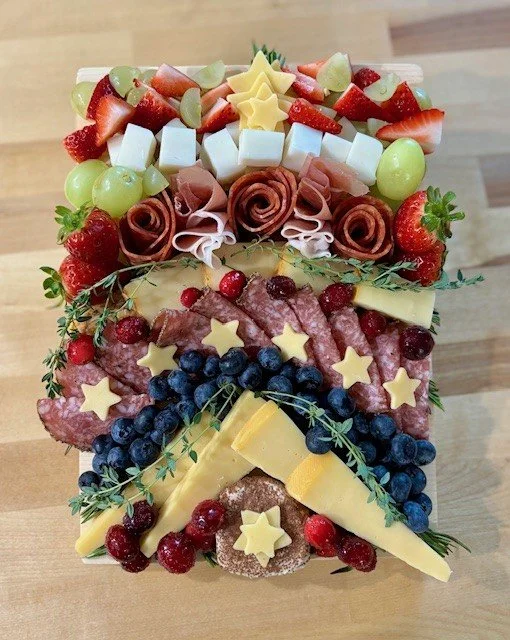 A festive charcuterie board decorated with strawberries, green grapes, cubes of cheese, star-shaped cheese slices, rolled ham and turkey, blueberries, red and black currants, and garnished with sprigs of herbs.