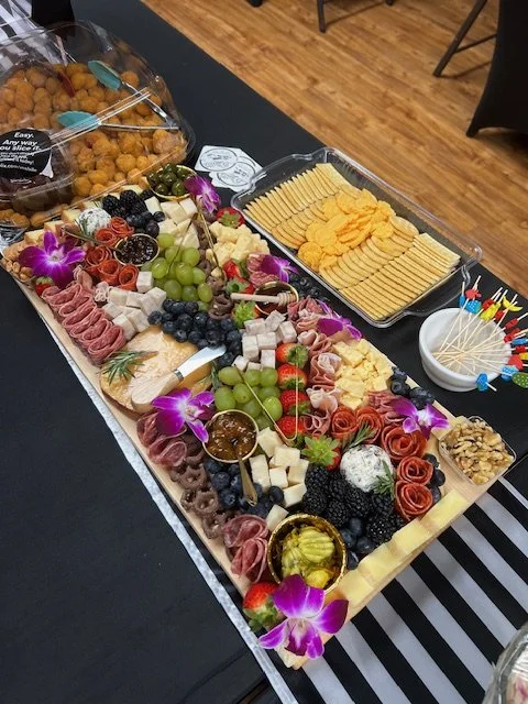 Charcuterie board with cheeses, grapes, berries, meats, nuts, and edible flowers, alongside crackers and dipping sauces on a black table.