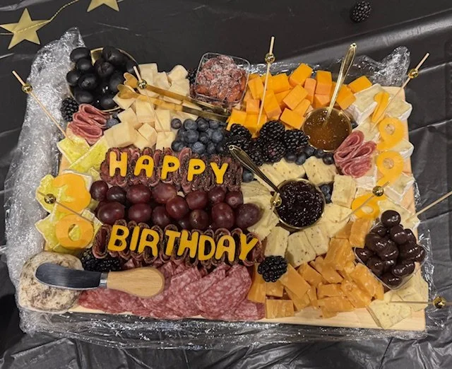 A large birthday cheese and fruit platter with assorted cheeses, grapes, blackberries, blueberries, cured meats, honey, and cheese markers that spell out "HAPPY BIRTHDAY."