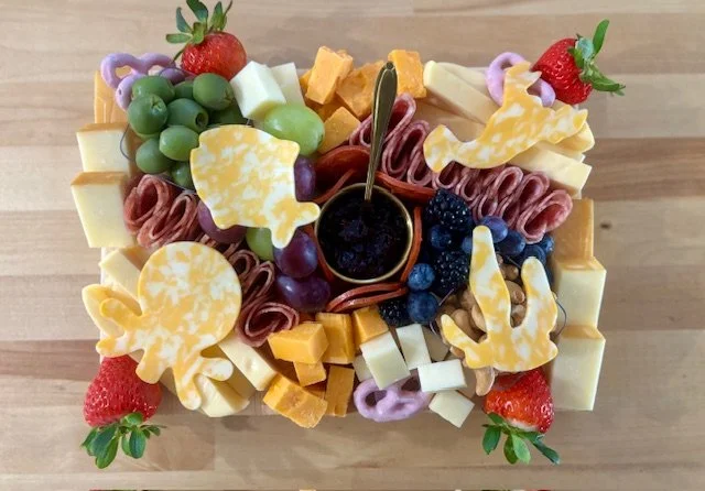 A cheese and fruit platter with strawberries, green grapes, blueberries, blackberries, cubed cheese, sliced meats, and yellow and purple decorative cheese slices, with a small bowl of dark dipping sauce in the center, arranged on a wooden board.