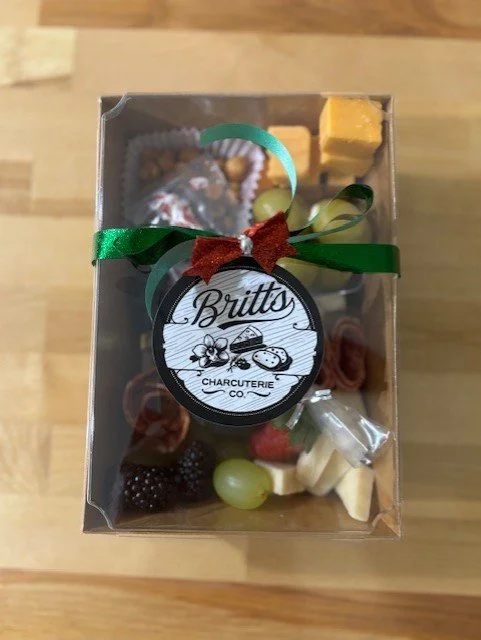 Gift box containing assorted chocolates, cheese, grapes, strawberries, and a label from Britt's Charcuterie Co. with a green ribbon and a red bow.