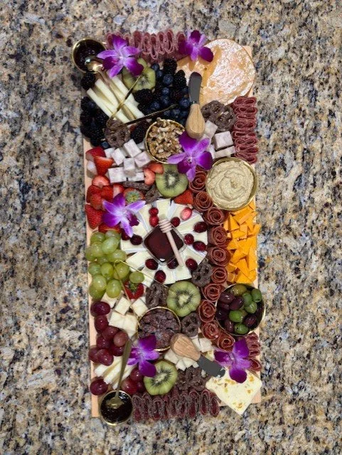 Assorted candies and fruits arranged on a rectangular platter with purple flowers for decoration.