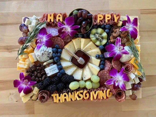 A large rectangular cheese and fruit platter decorated with purple orchids, grapes, blueberries, blackberries, and assorted cheeses, featuring "Happy Thanksgiving" in orange letters.