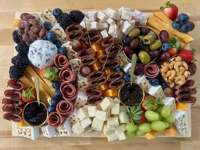 A large cheese and fruit platter with assorted cheeses, grapes, strawberries, blueberries, blackberries, honey, cashews, and slices of cured meats.
