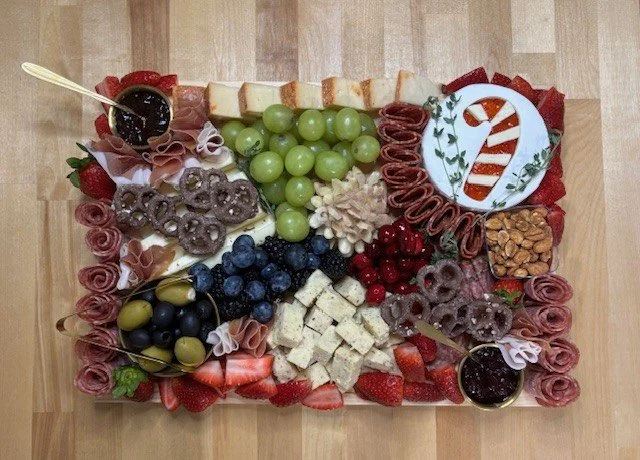 A festive appetizer platter with cheese cubes, green grapes, strawberries, blueberries, red grapes, sliced meats, pretzels, crackers, and two small bowls of jam, all arranged on a rectangular tray decorated for a holiday celebration.