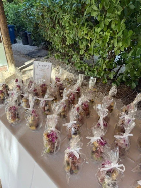A table with small clear plastic bags each containing a dessert, tied with white ribbons, arranged outdoors near green bushes.