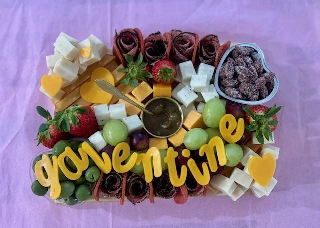 A colorful cheese and fruit platter with strawberries, grapes, cheese cubes, heart-shaped cheese, rolled meats, a small container of dried berries, and decorative yellow text spelling "love" on a pink paper background.
