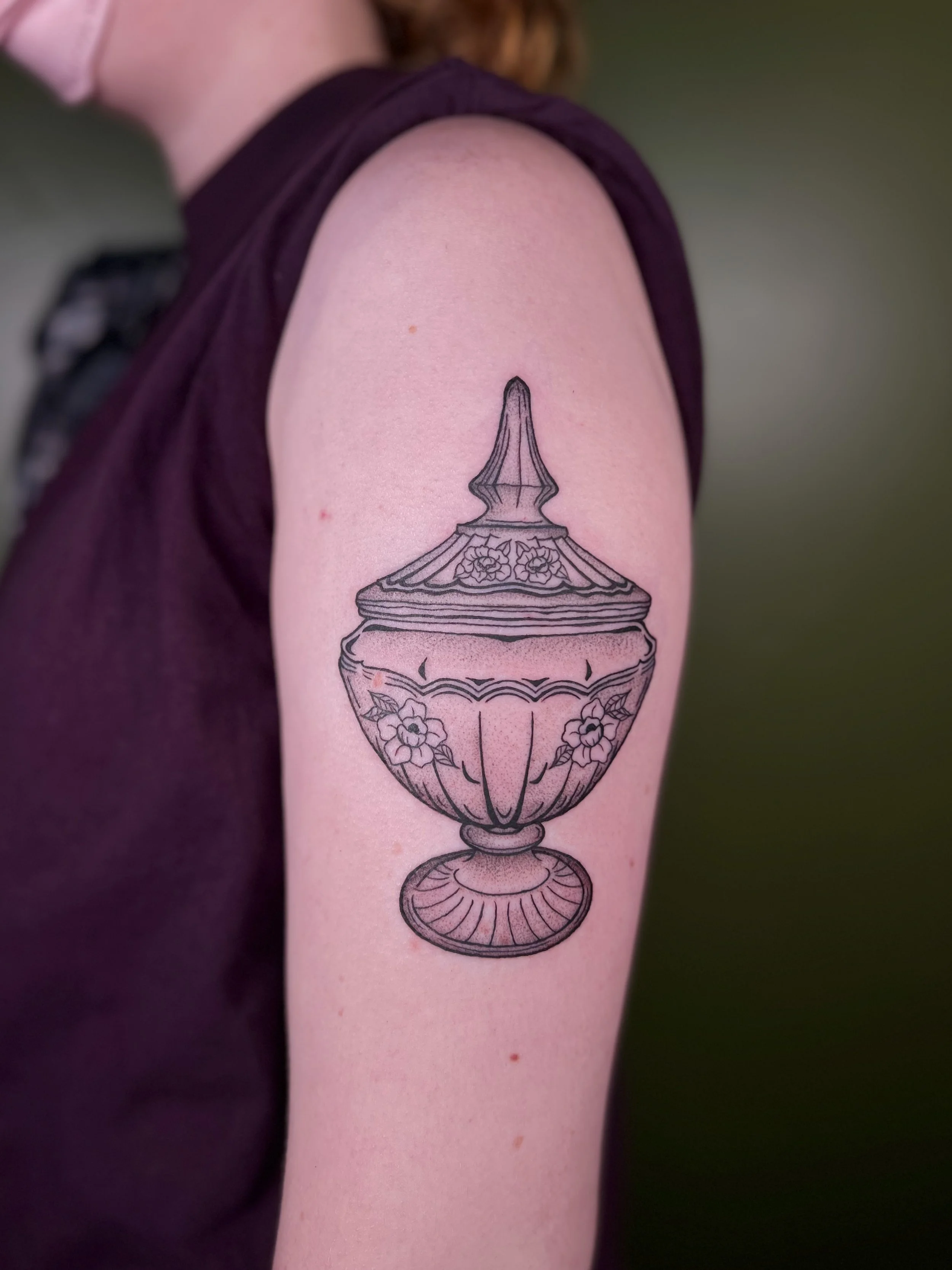 Black outline tattoo of an ornate jewelry box with floral details on the arm of a person wearing a sleeveless dark top.