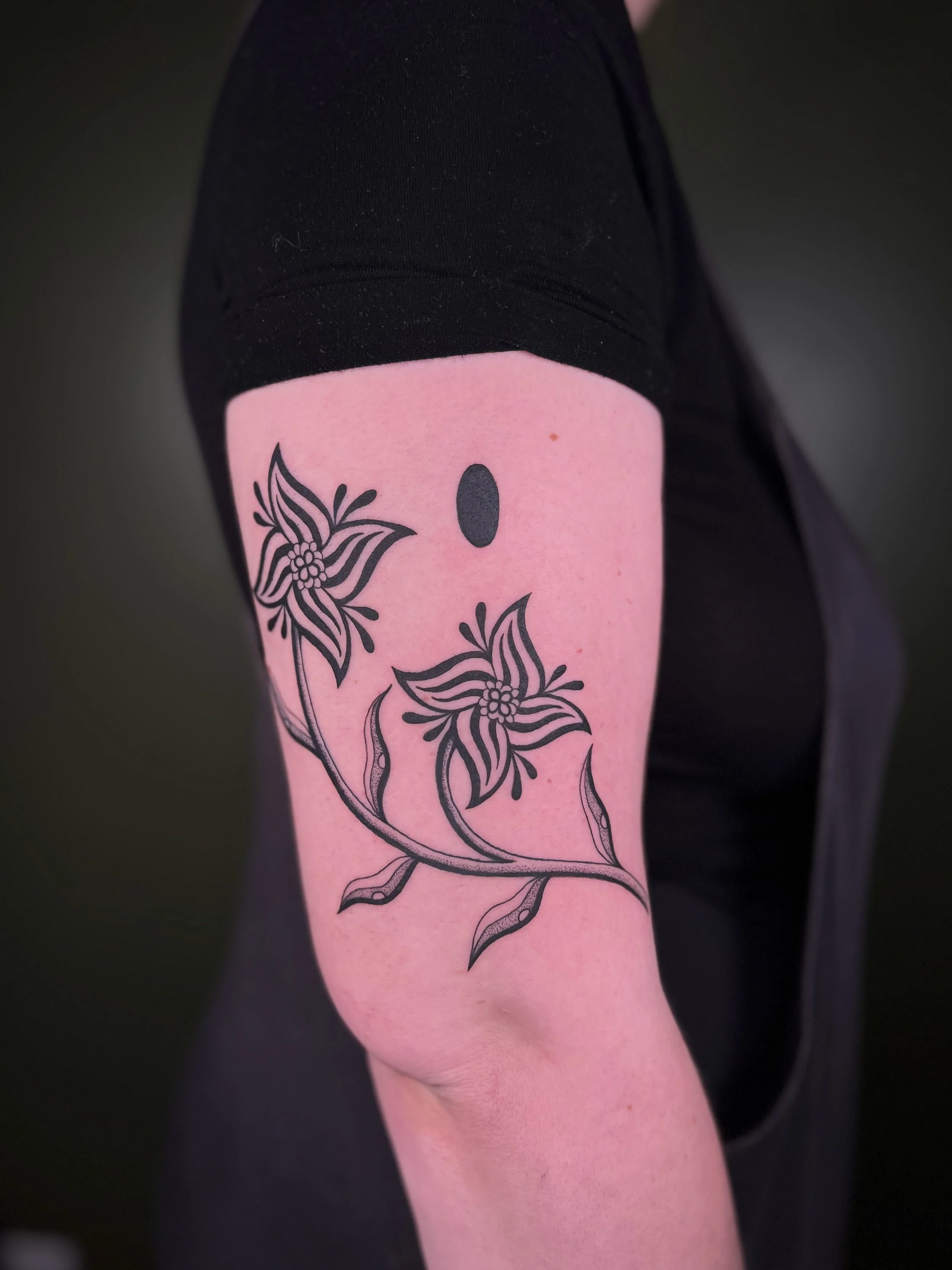Tattoo of a flower with two blossoms and a small oval above on a person's upper arm.