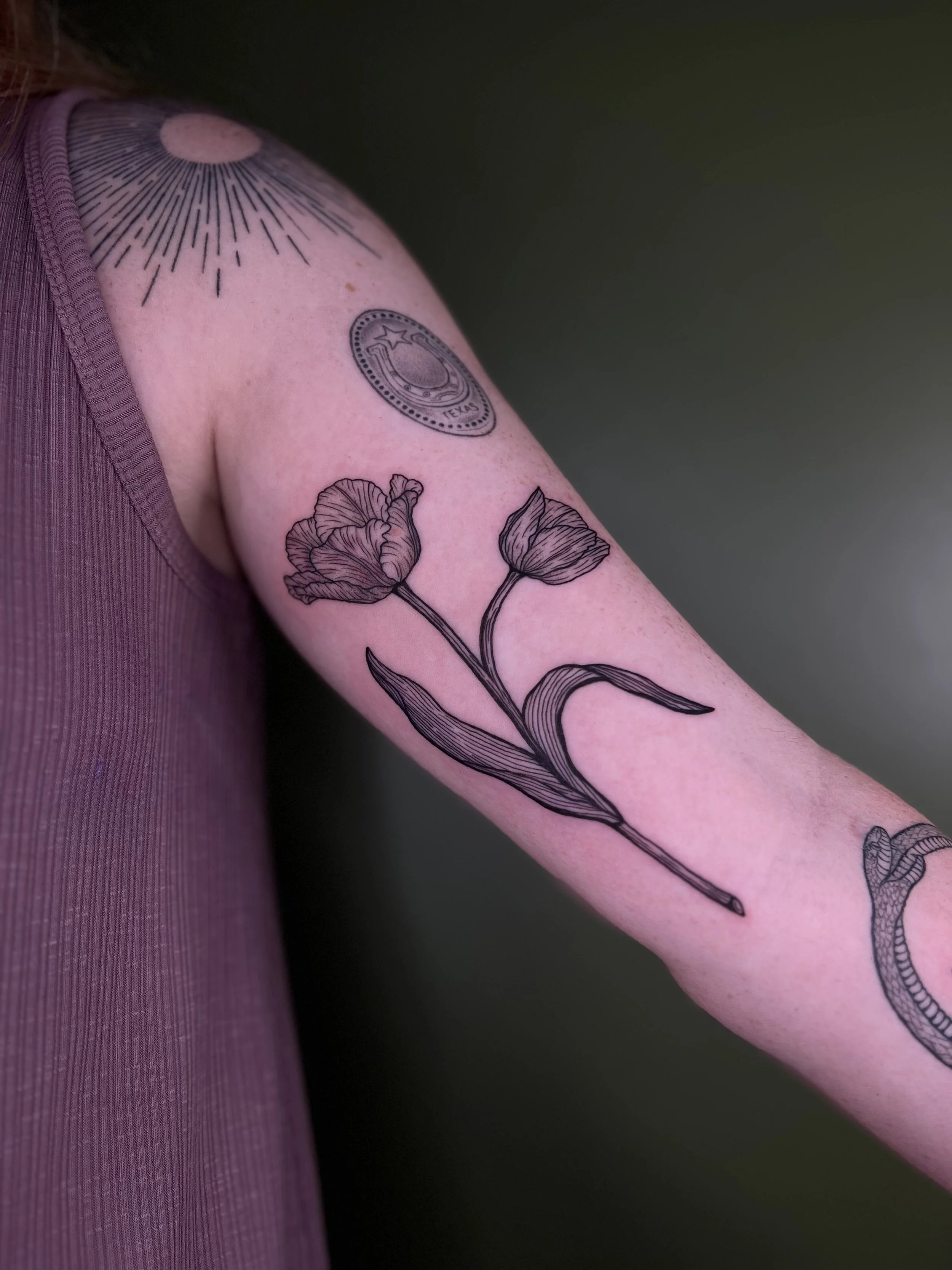 Close-up of a person's arm with a floral tattoo of two poppy flowers, an upper shoulder tattoo with a circular design, and other tattoos partially visible.