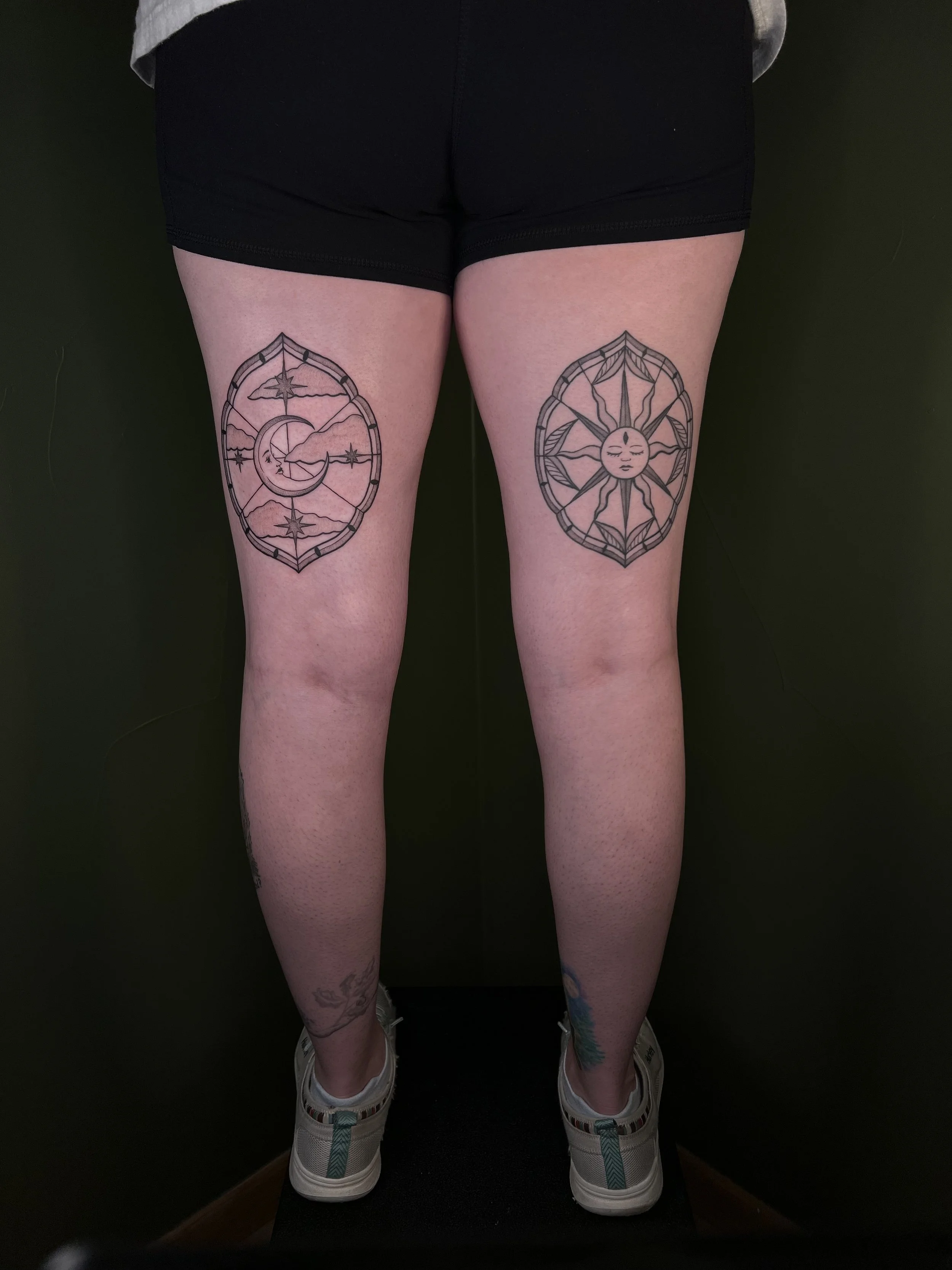 Tattoo of a moon and sky with clouds on the left thigh, and a sun with rays and face on the right thigh, both within circular frames.