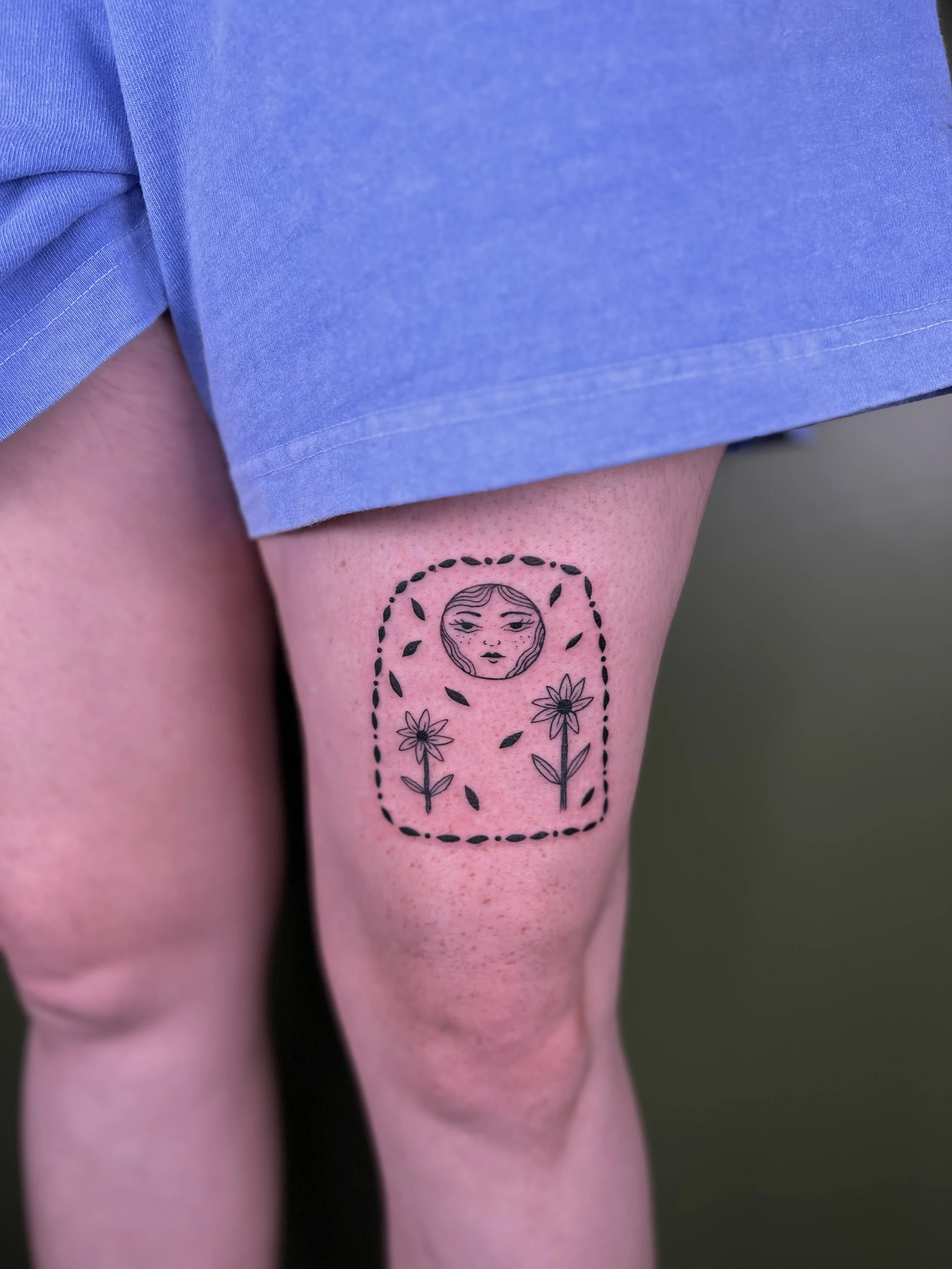Tattoo of a woman's face within a circle, surrounded by two flowers and a dashed border, on a person's thigh.