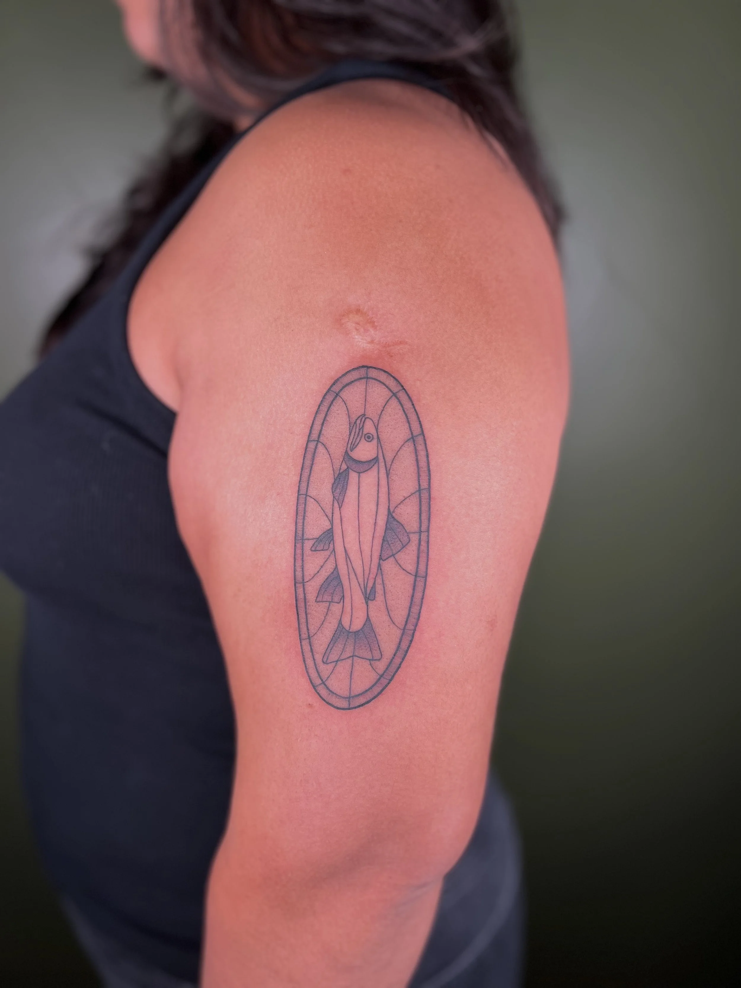 Tattoo of a fish inside an oval on a person's upper arm.