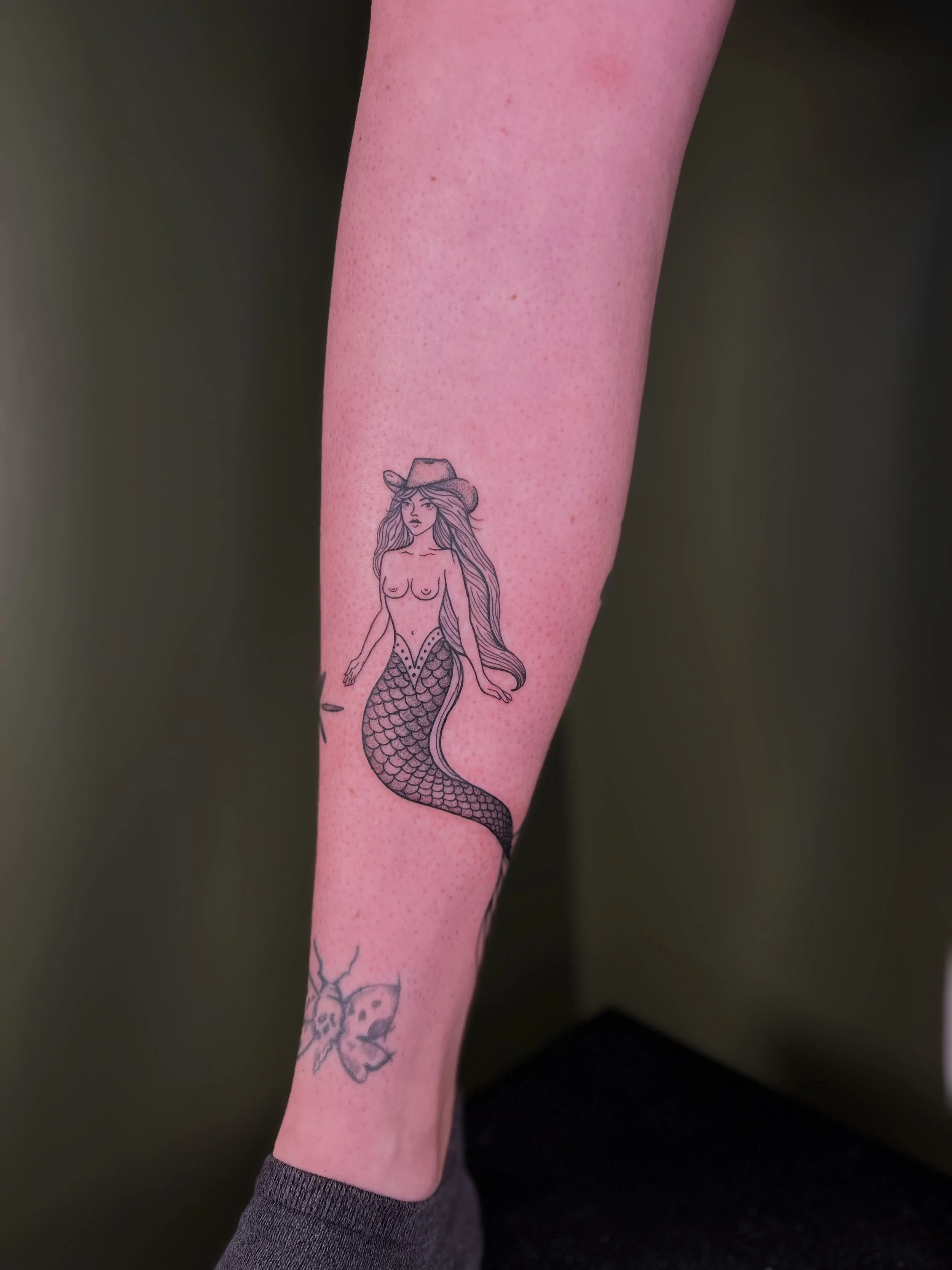 Tattoo of a mermaid with long hair, wearing a hat, on a person's lower leg.