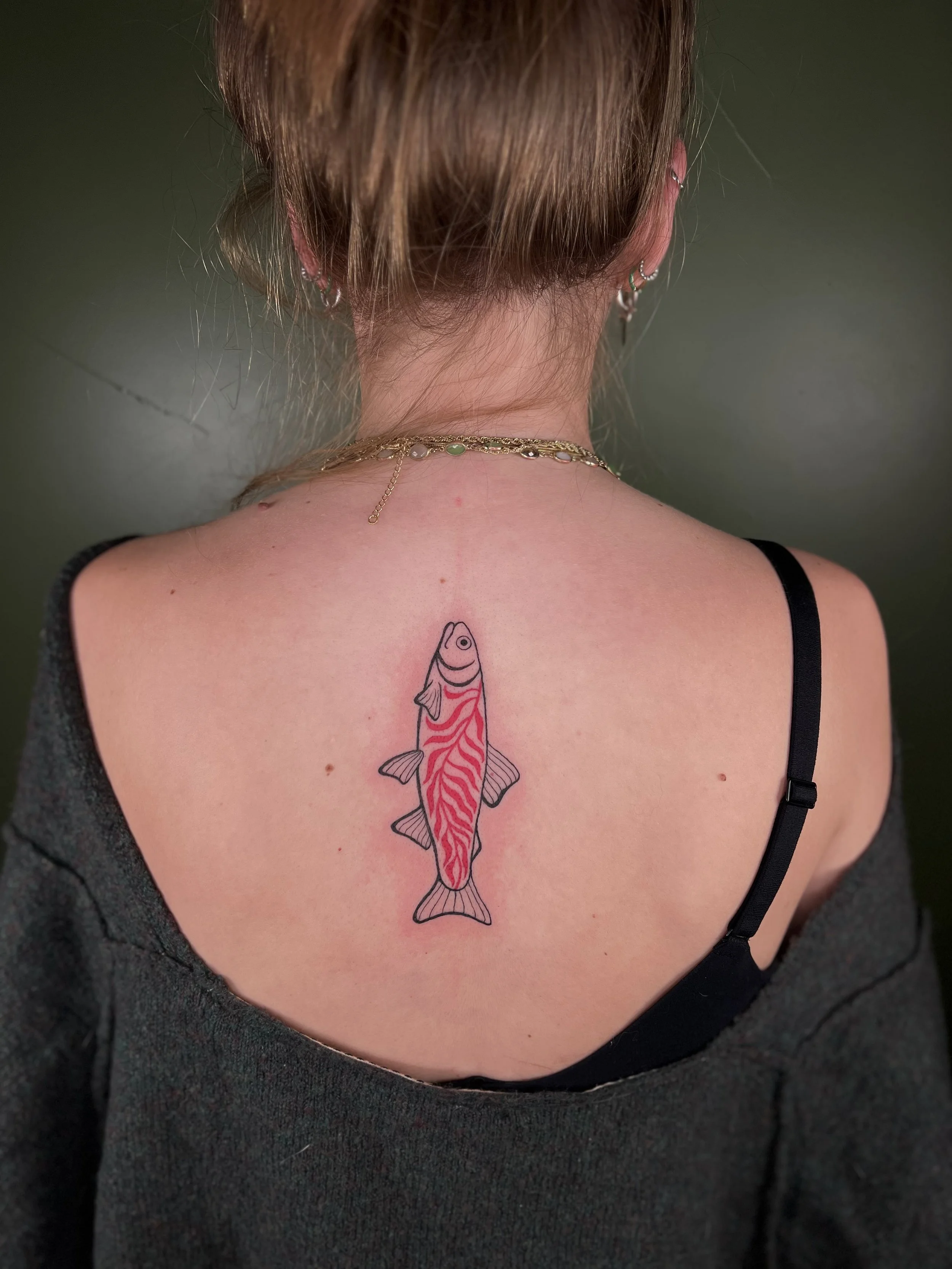 Back of woman with a tattoo of a fish on her upper back, wearing a black strap top and gold jewelry, against a green background.