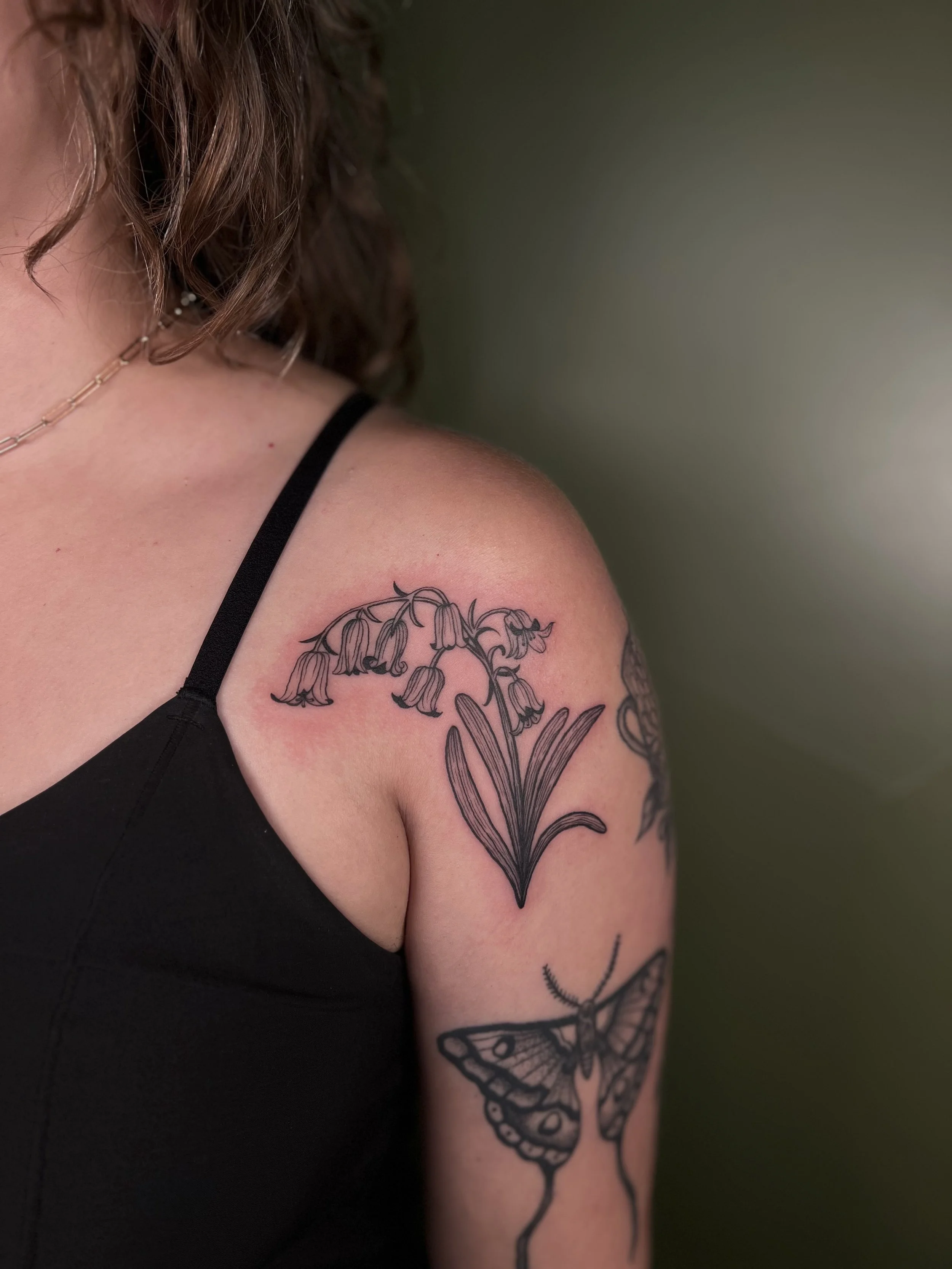 Tattoo of a bell-shaped flower with long leaves on shoulder, and a moth underneath it on arm, all in black ink.